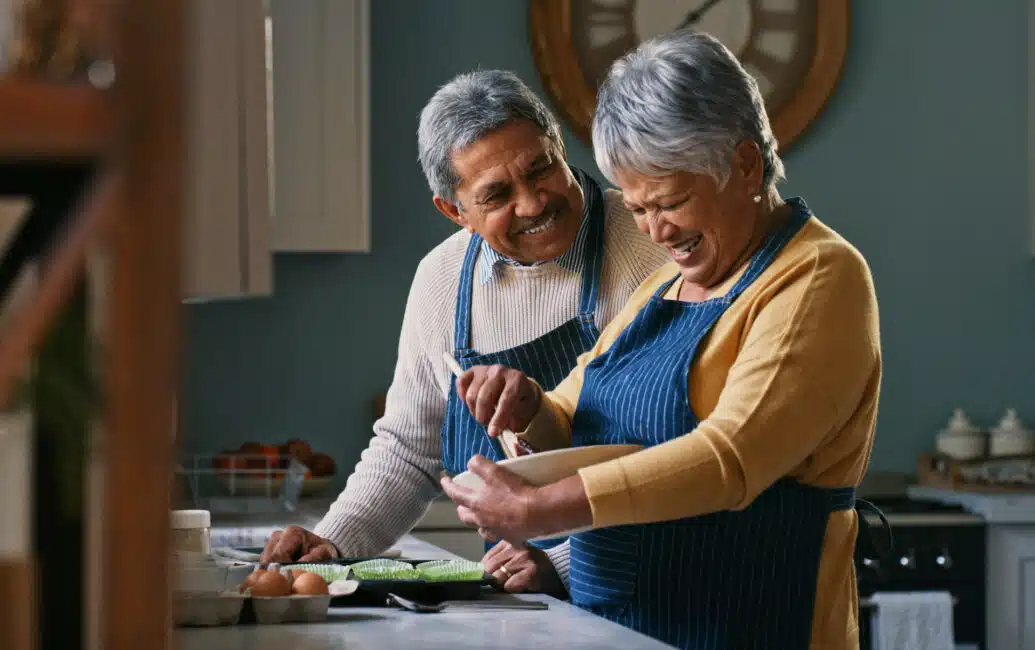 Seniors wearing aprons in a kitchen, woman is mixing in a bowl while man looks on close beside her
