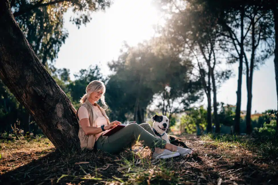 Fashionable senior woman with pet dog sitting under tree in forest park and reading book